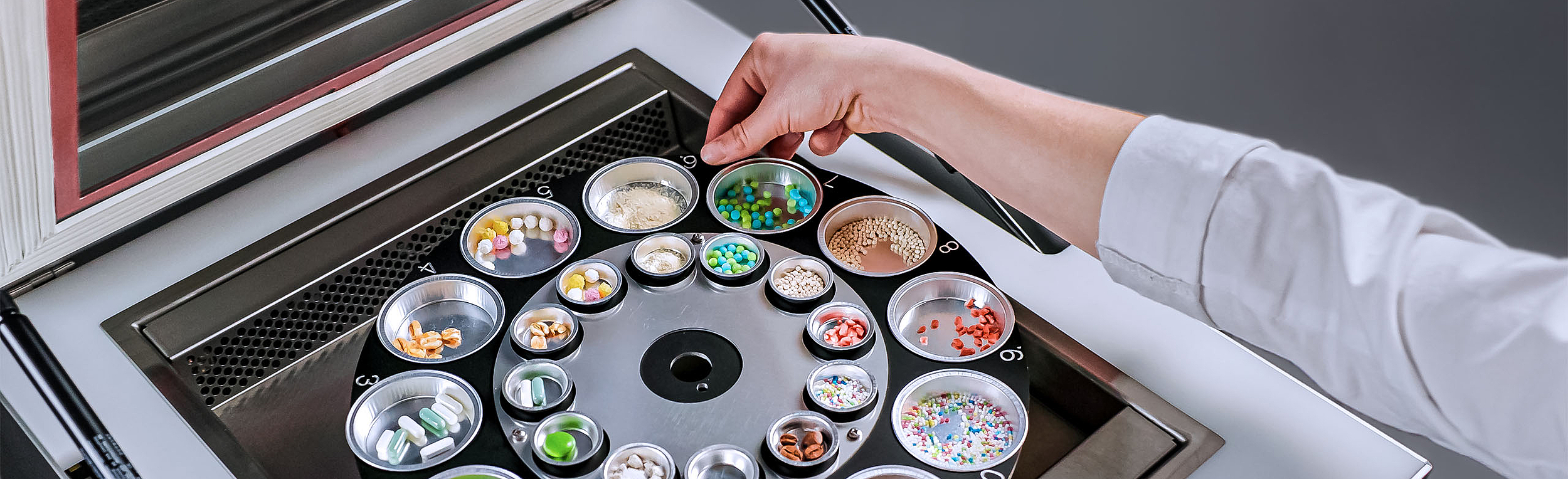 Nahaufnahme einer Person, die ein SPS bedient und verschiedene kleine Behälter mit Pillen, Süßigkeiten, Streuseln und anderen Materialien handhabt. 
