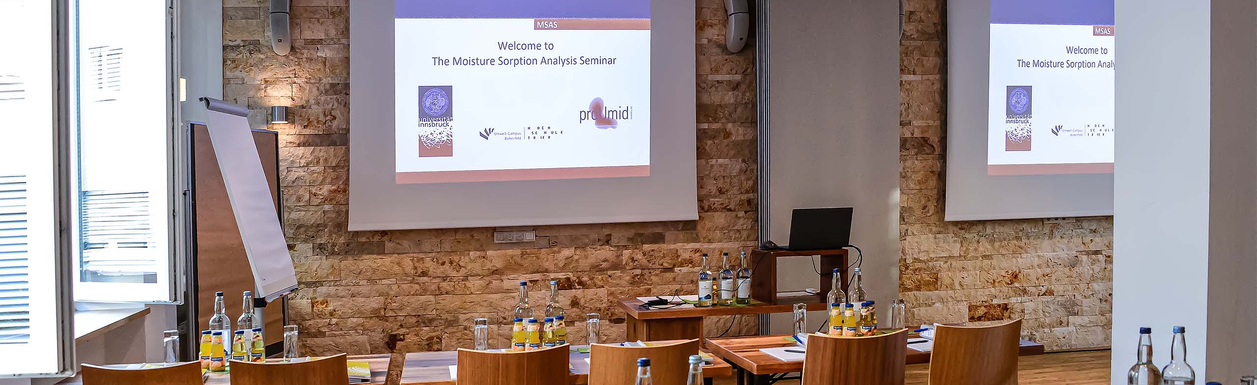 Well-equipped proUmid seminar room with wooden tables and chairs, a flipchart, refreshments, and projection screens for the 'Moisture Sorption Analysis Seminar'. The presentation displays the proUmid logo and welcome text. Ideal for training and workshops.